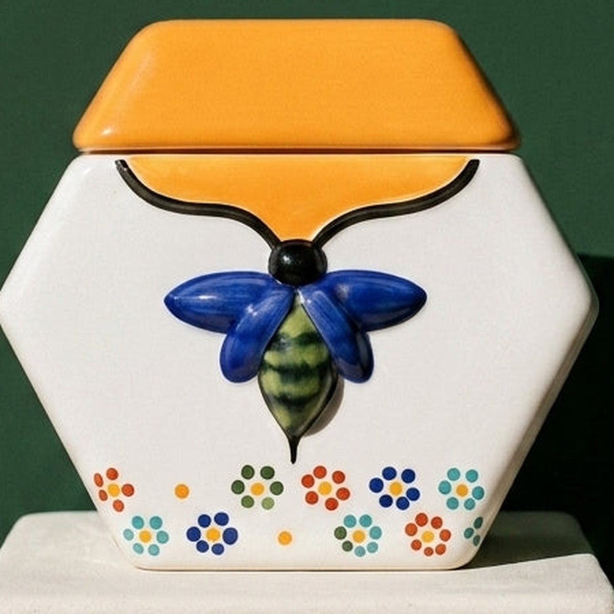 Close-up of the blue bee in relief and hand-painted dot flowers on the De Todas Las Flores ceramic vessel, against a deep green wall.